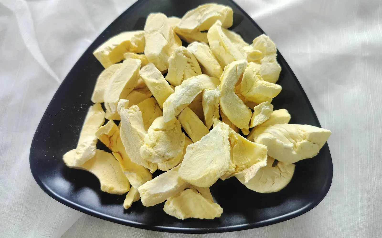 Black triangular plate heaped with pale yellow freeze-dried durian pieces on white fabric.