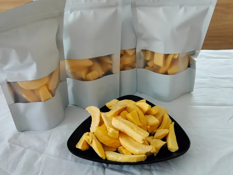 Three silver stand-up pouches with clear windows showing dried mango, with a black plate of mango strips in front.
