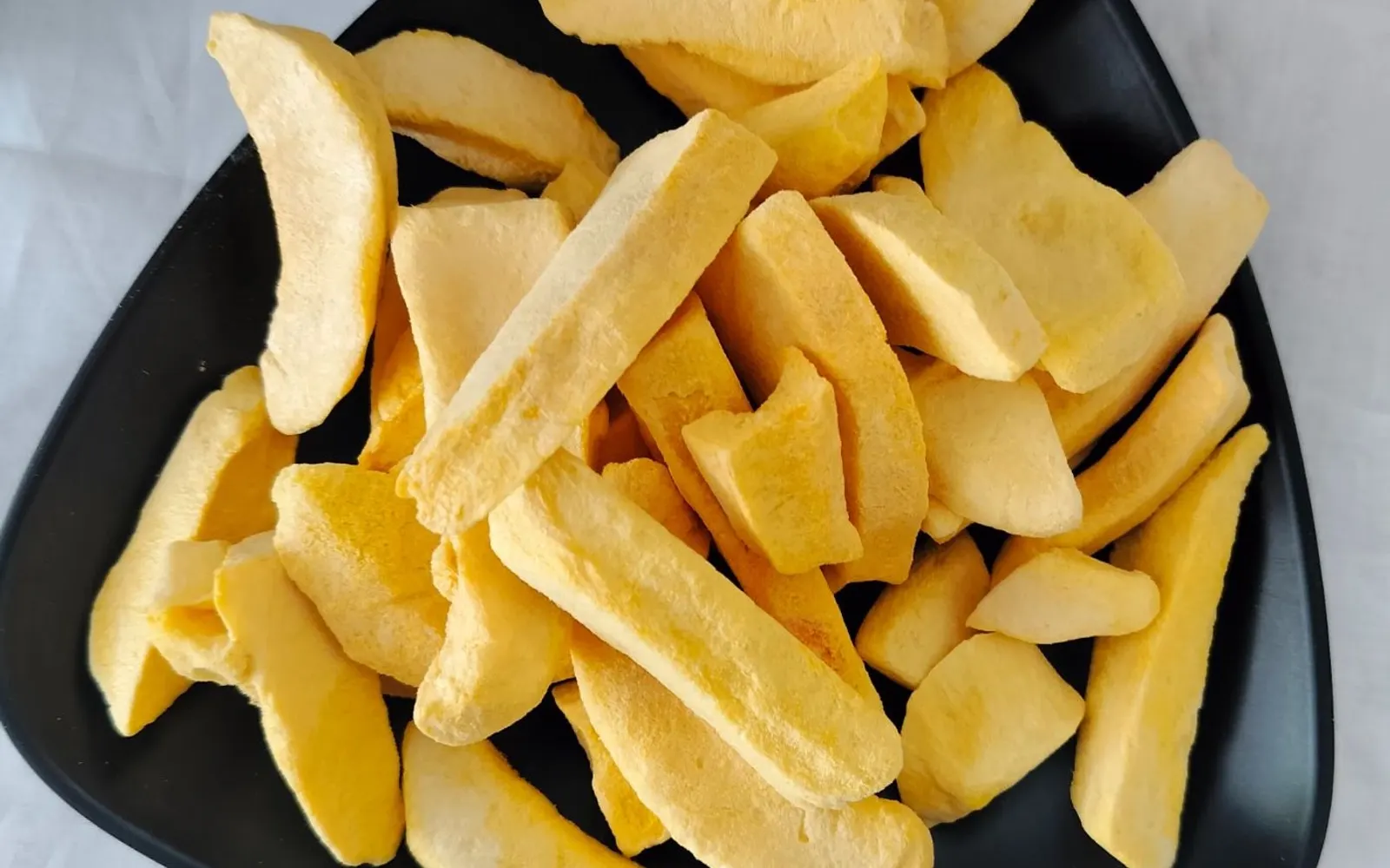 Black plate of pale, porous freeze-dried mango sticks and wedges.