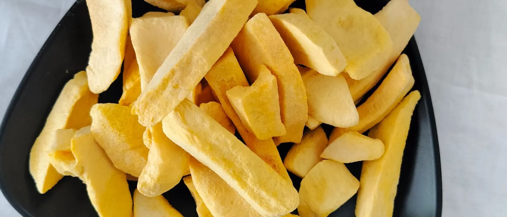Black triangular plate of golden freeze-dried mango sticks on a white cloth.
