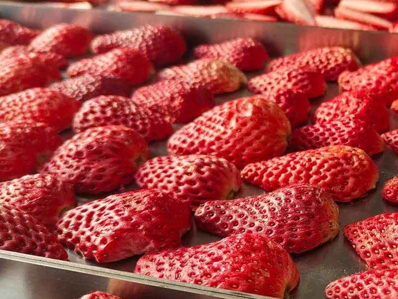 Strawberries arranged on metal trays in a factory, sliced strawberries on trays behind.