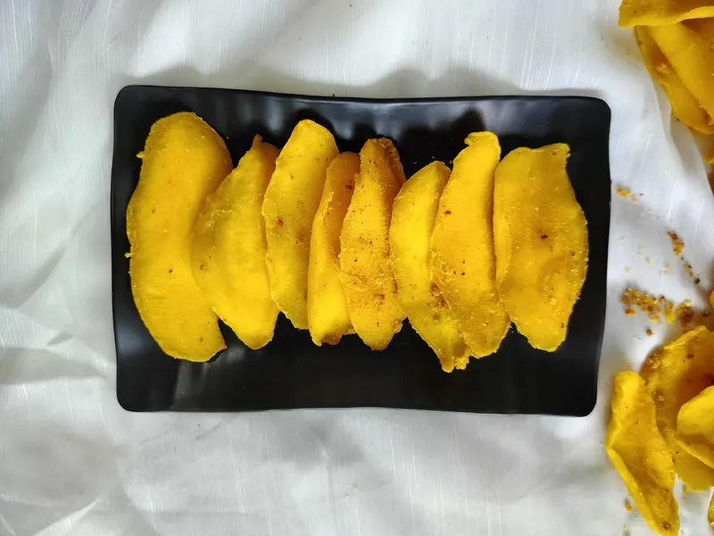 Black rectangular plate with a row of chilli-dusted soft-dried mango slices on white cloth.