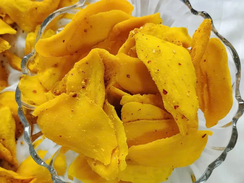 Footed glass bowl filled with chilli-dusted soft-dried mango slices on a white surface.
