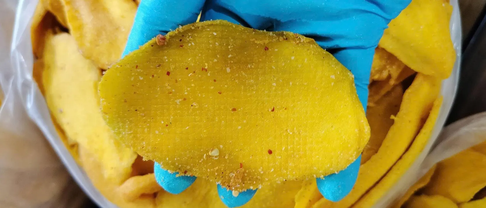 Gloved hand holding a chilli-dusted soft-dried mango slice above a tray of dried mango.