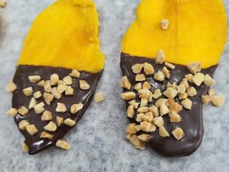 Close-up of two mango slices half-dipped in dark chocolate and sprinkled with chopped peanuts.