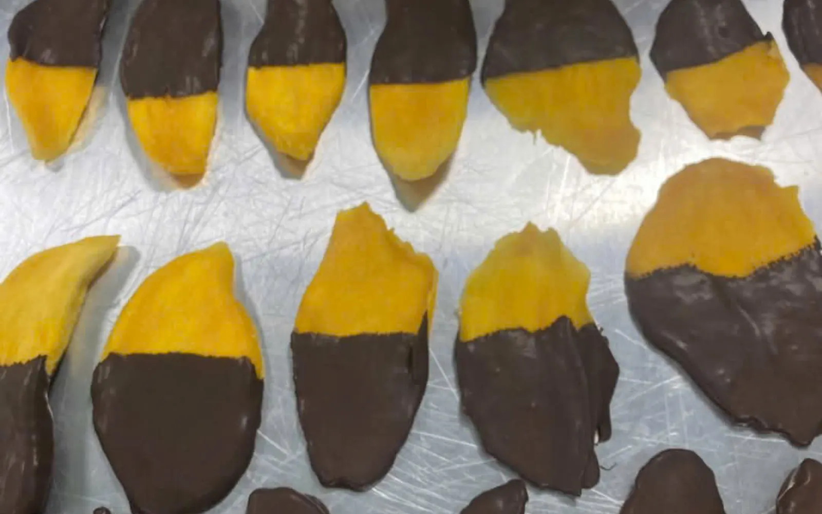 Neat rows of dried mango slices half-dipped in dark chocolate on a stainless tray.