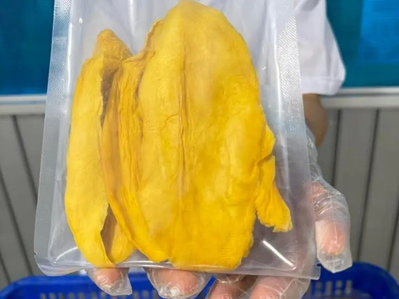 Worker in a white coat holding up a large vacuum pack of dried mango slices above a blue crate.