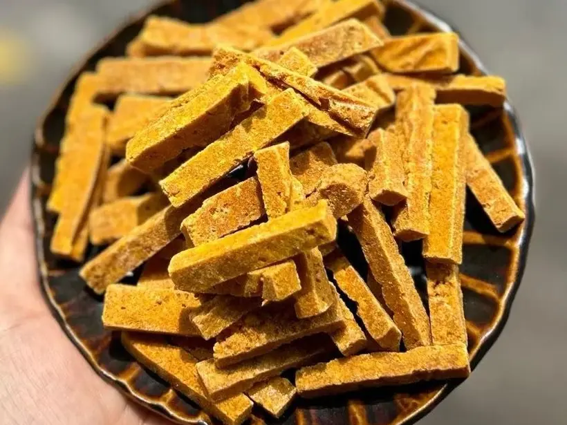 Hand holding a dark ceramic plate of orange-yellow dried ginger sticks.
