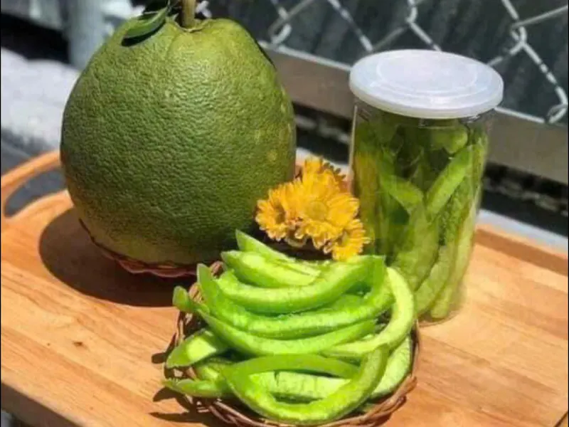 Whole green pomelo on a wooden board with a basket of fresh peel strips and a jar of preserved peel.