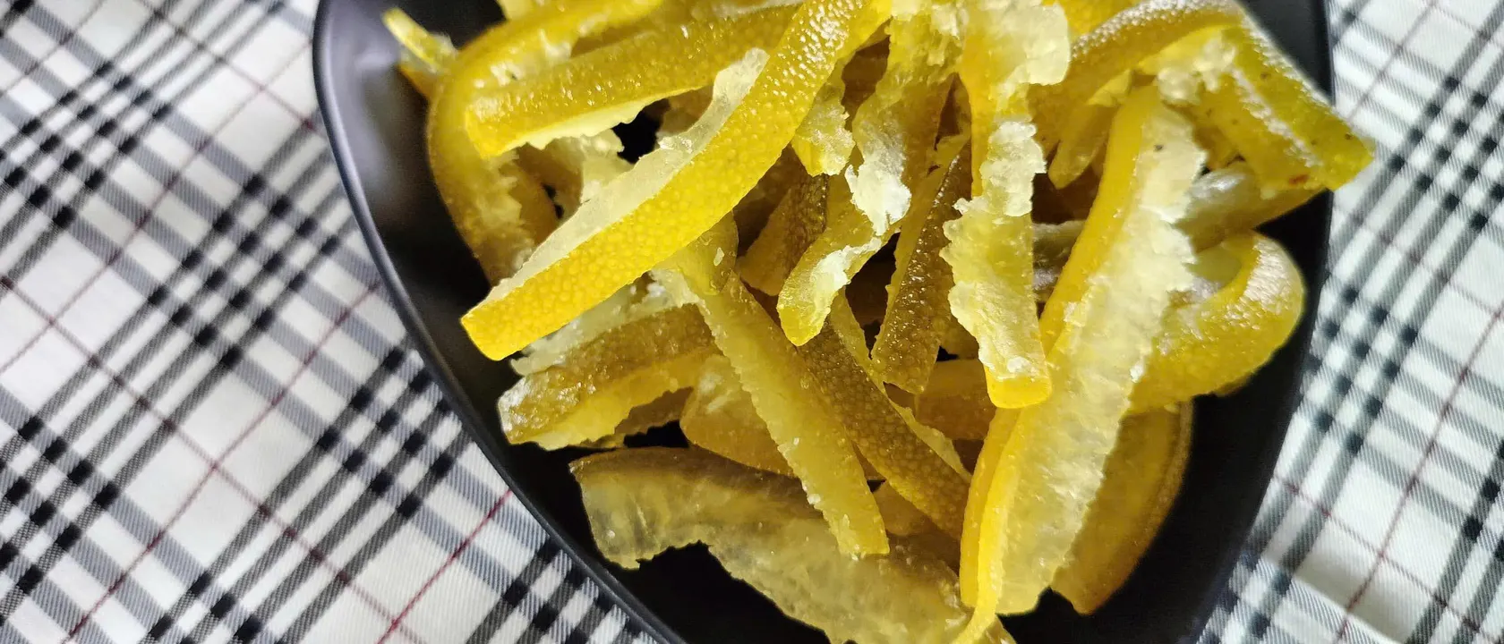 Close-up of a black plate heaped with dried pomelo peel strips on a checked cloth.