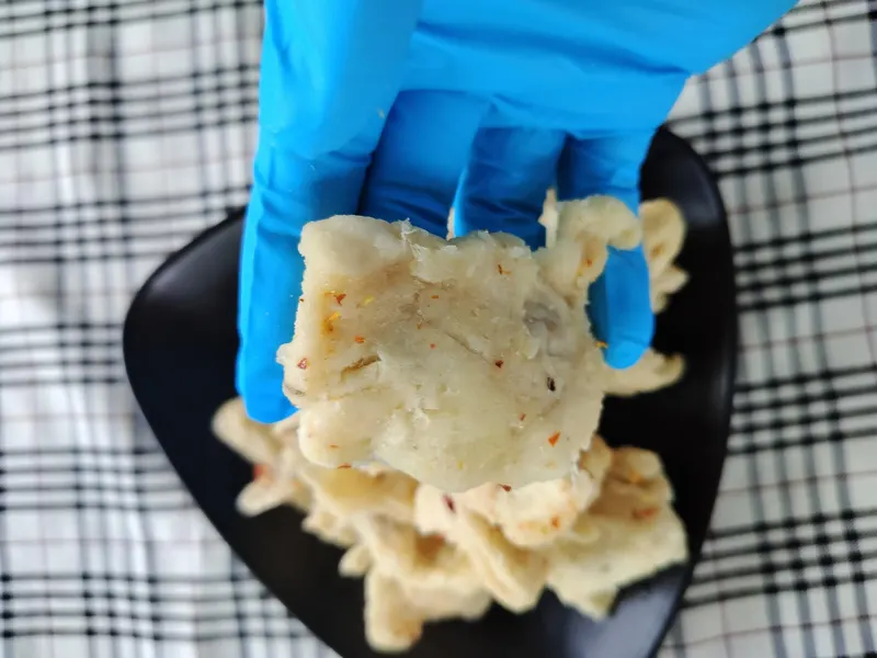 Gloved hand holding a chilli-flecked soft-dried soursop piece above a black plate.