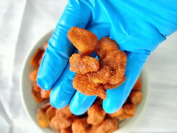 Gloved hand scooping soft-dried strawberries above a white bowl of the product.